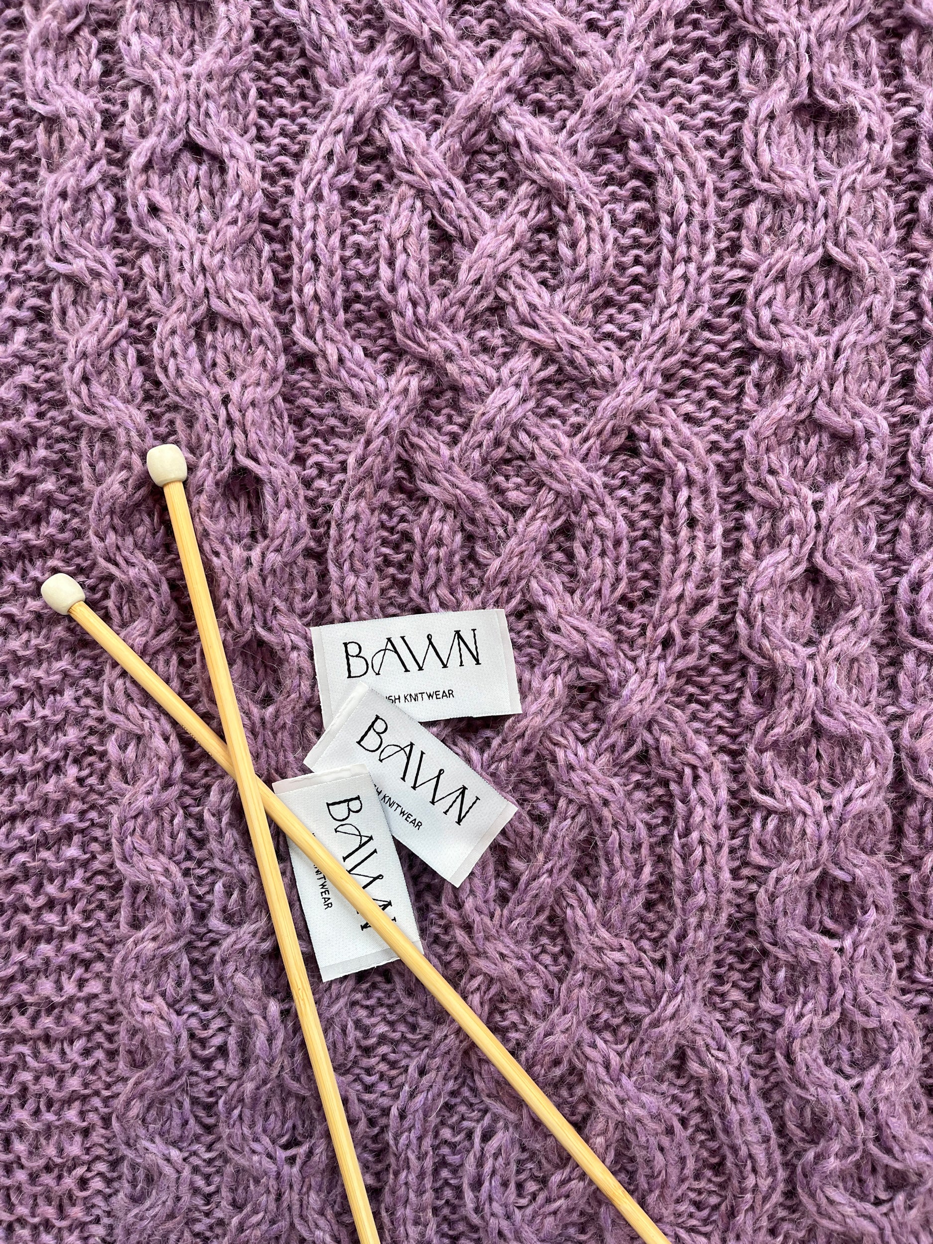 Flat laid hand knit pink cable pattern with two knitting needles and BAWN back of neck labels. 